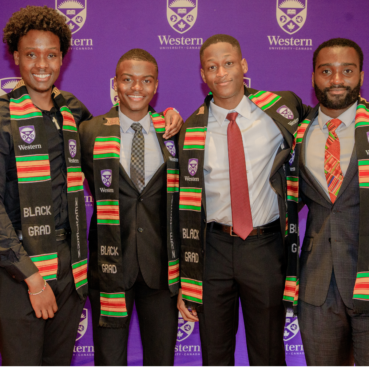 Four Black graduates stand for a photo in front of the Western purple backdrop