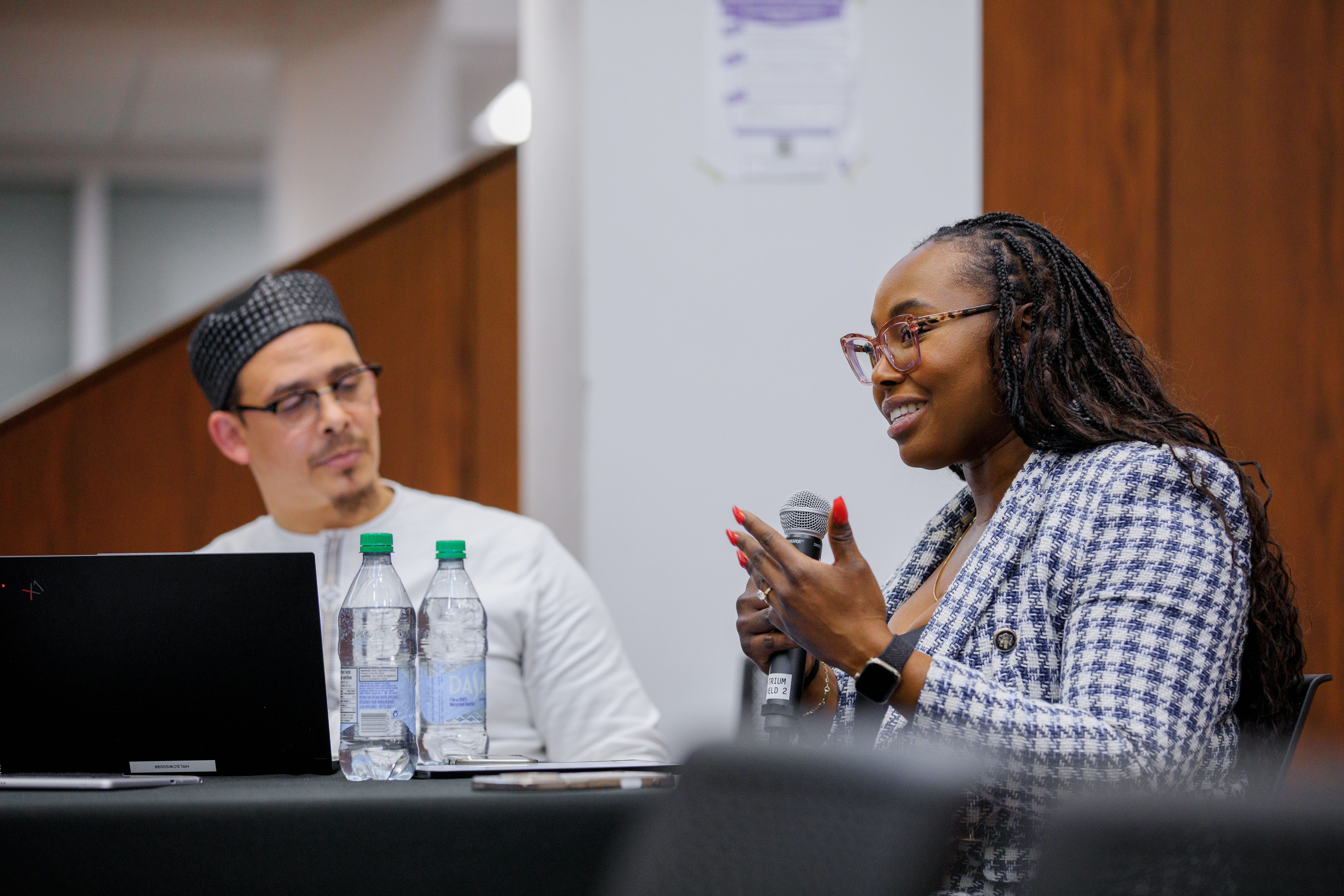 This is a photo of the Black Resistance Against the Status Quo Panel event on February 24, 2025. Dr. Remi Warner, Interim Executive Director of the Human Rights Legal Support Centre, listens to Tega Odjevwedje, Co-president of the Black law Student's Association, Western Chapter. Tega is wearing a patterned blazer and is speaking into the microphone and gesturing with her hand. 