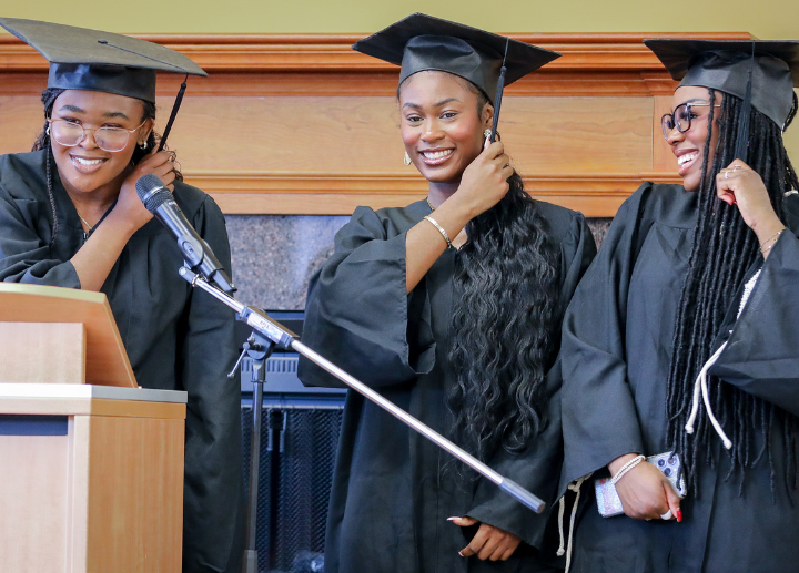 Three Black students wear graduation caps and gowns and stand at a podium.