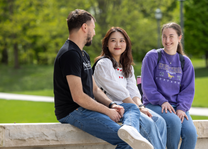 Three students sit outside during the summer on campus and smile