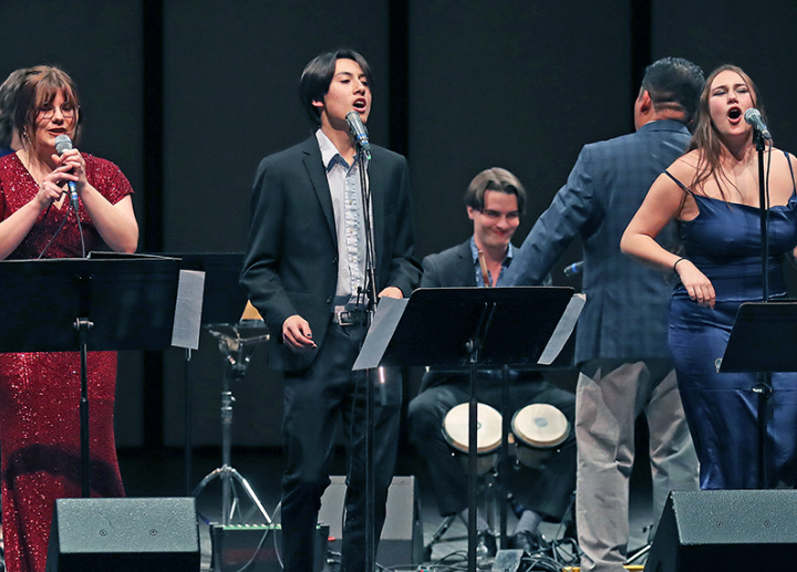 Three performers wearing formal outfits stand on a dark stage, singing with a band in the background.