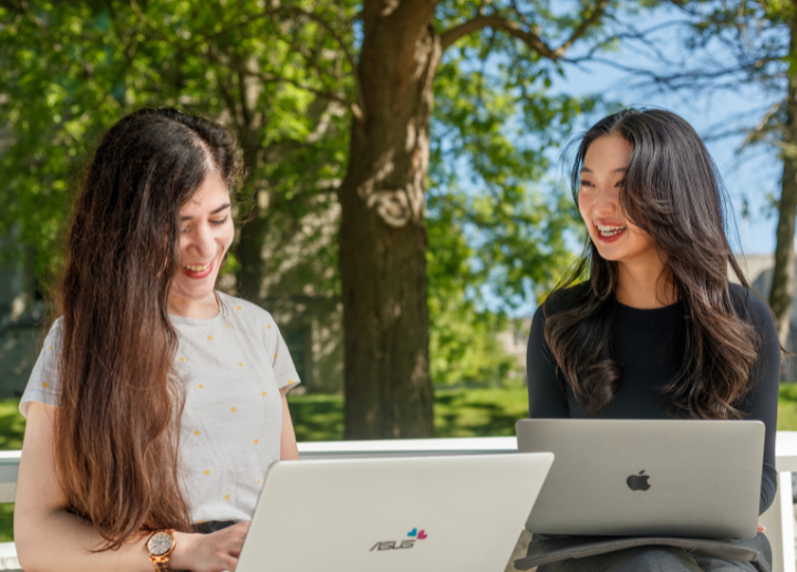 Two students smiling, working on their laptops outside on campus